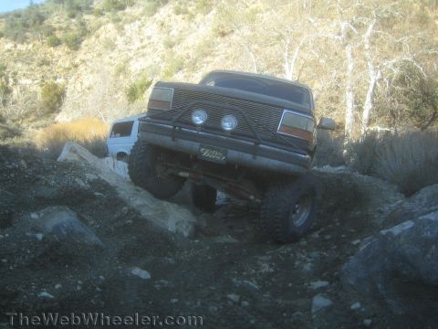 1990 Ford Bronco - lifted dark Ford Bronco with round driving lights flexes over rocks on a hillside trail, a white-topped SUV behind it in a canyon.