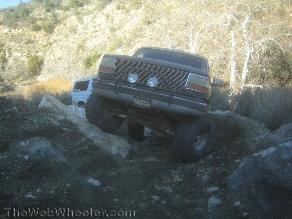 Lifted dark Ford Bronco with driving lights crawling rocks beside a tan SUV