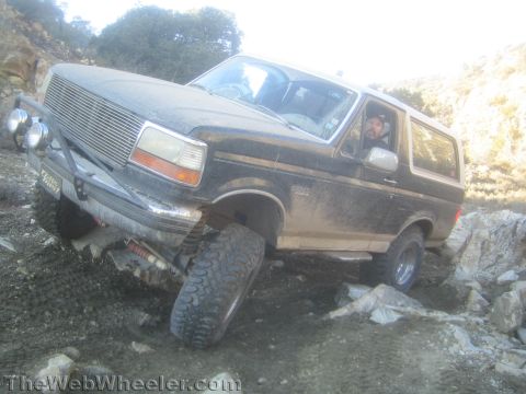1990 Ford Bronco - heavily mud-splattered lifted Ford Bronco with billet grille and driving lights crawls over rocks, the driver visible through the window.
