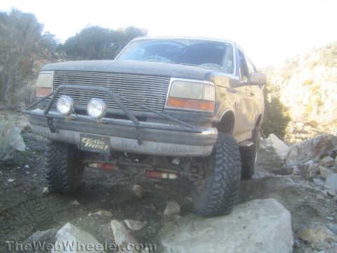 1990 Ford Bronco - Low front view of a dusty lifted Ford Bronco with billet grille, brush guard and round driving lights crawling a rocky trail.