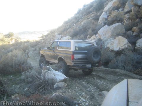 1990 Ford Bronco - lifted two-tone Ford Bronco with a rear-mounted spare tire crawls up a rocky desert trail beside a steep rock face.