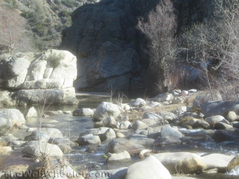 1990 Ford Bronco - shallow rocky creek strewn with large boulders winds past a sunlit cliff face in a dry canyon.