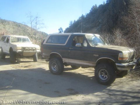 1990 Ford Bronco - lifted dark two-tone Ford Bronco parked on a dirt clearing next to a white off-road SUV in a hilly area.