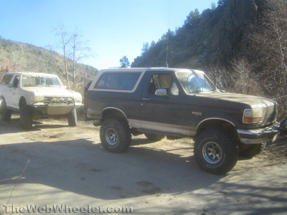 Lifted dark Ford Bronco and a white SUV parked on a dirt clearing in hills