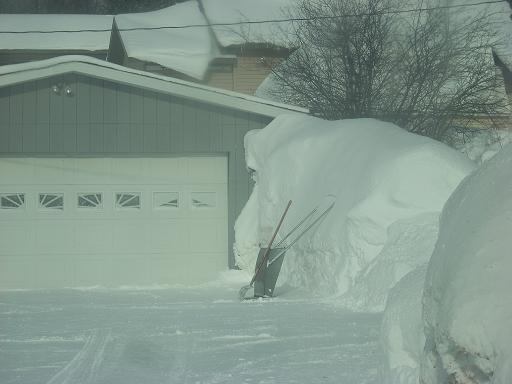 Snow-covered garage with a large snowdrift and shovels, tire tracks in snow