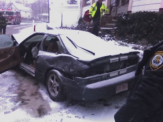 Wrecked dark gray hatchback coupe in snow at a crash scene with fire truck behind