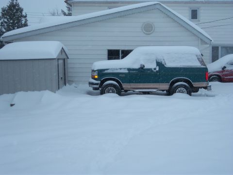 1996 Ford Bronco - A green 1996 Ford Bronco is partially buried in snow next to a house and shed, with another vehicle visible in the background.