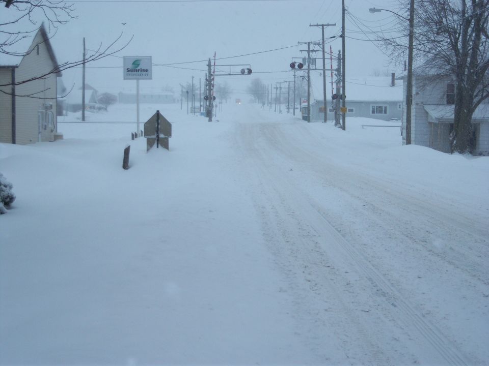 Snow-covered street with houses, power lines, and a sign visible