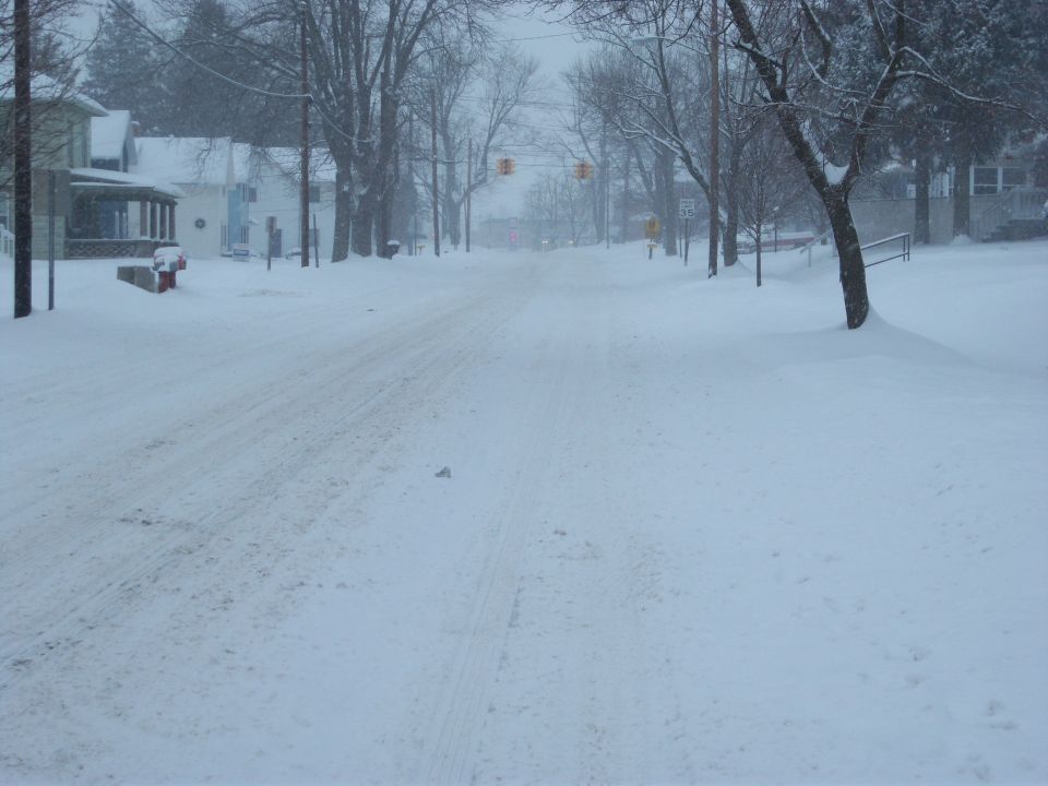snowy street with houses and trees, traffic lights visible