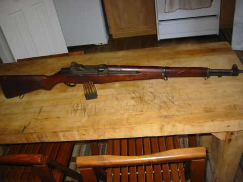 1996 Ford Bronco - A vintage M1 Garand rifle with a loaded clip rests on a worn wooden table, with chairs visible below.