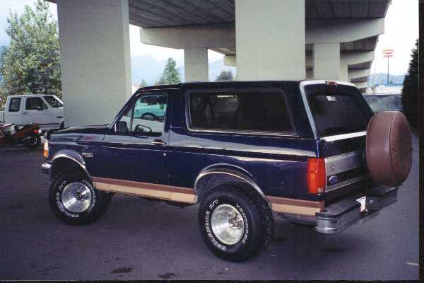Dark blue and tan Ford Bronco SUV with rear spare tire cover, parked under a highway overpass, rear driver-side view