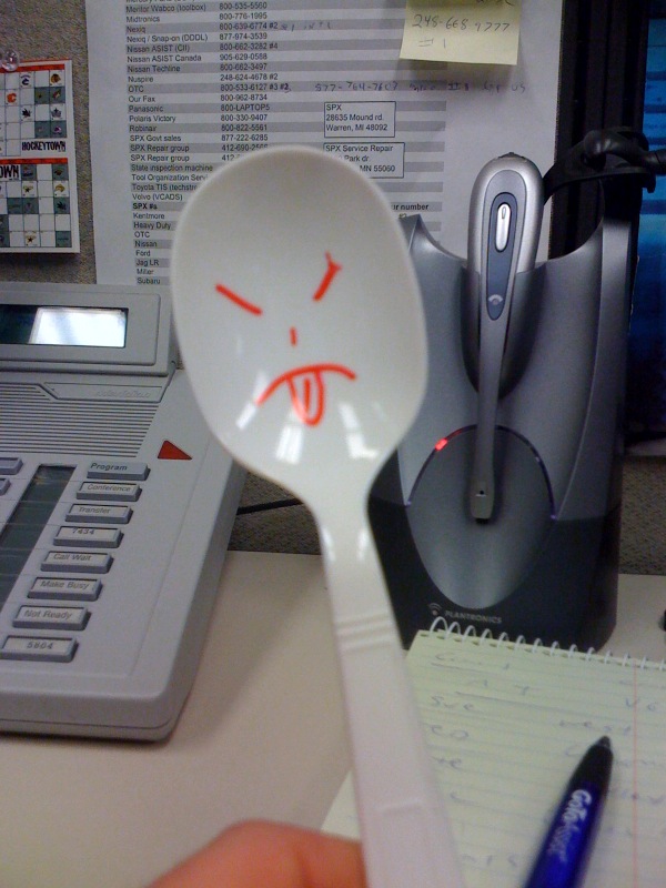 Spoon with a grumpy face drawn on it, held in front of a desk phone and headset