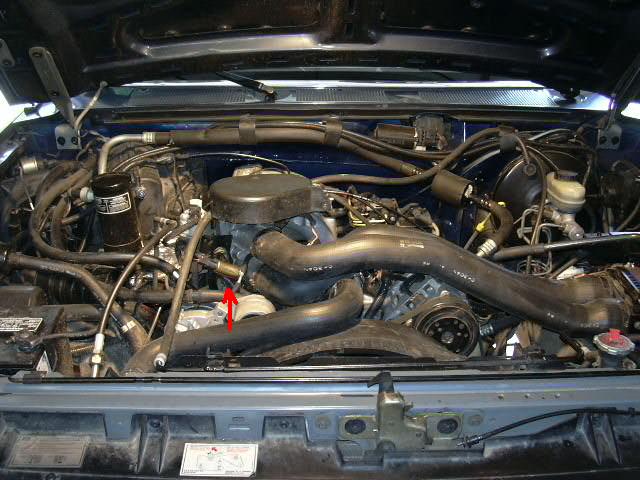 Engine bay with hoses, belts, and engine components visible. Red arrow points to a hose connection