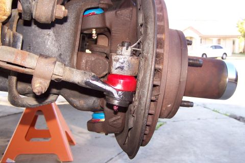 1990 Ford Bronco - Close-up of a vehicle's suspension components, including a ball joint and steering linkage, with a house and car visible in the distance.