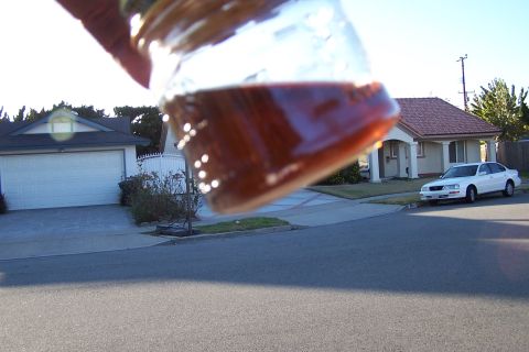 1990 Ford Bronco - A jar of amber liquid is held up, obscuring part of a suburban scene with a house and a white car.