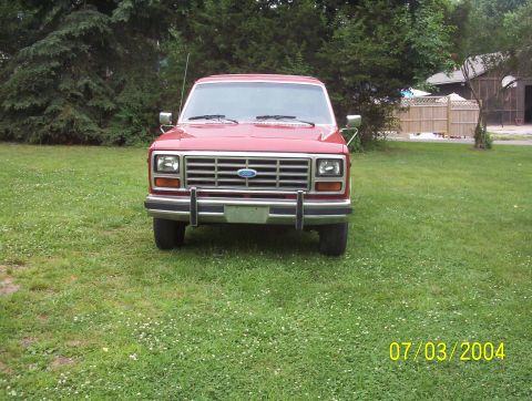 1985 Ford Bronco - A red Ford Bronco is shown from the front, parked on a grassy lawn with trees in the background.