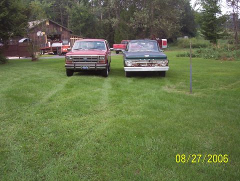 1985 Ford Bronco - Two Ford vehicles, a Bronco and an F-100 truck, are parked on a grassy lawn with a barn in the background.