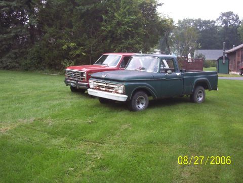 1985 Ford Bronco - A green 1965 Ford F-100 pickup truck is parked on the grass next to a red Ford truck, with a house and trees in the background.