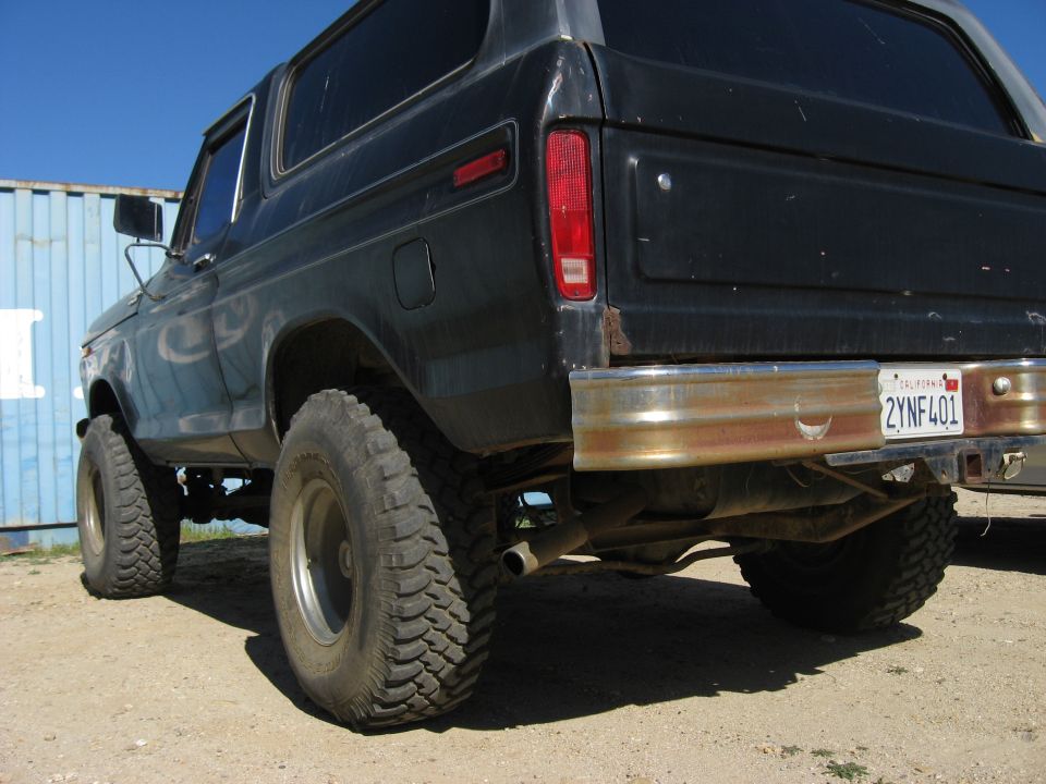 Rear view of a dark-colored Ford Bronco with large tires and a rusty bumper. California license plate reads 2YNF401