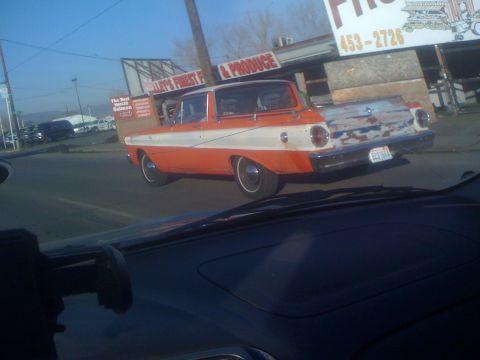 1983 Ford Bronco - An orange and white car drives down the road, its rear end showing rust and peeling paint.