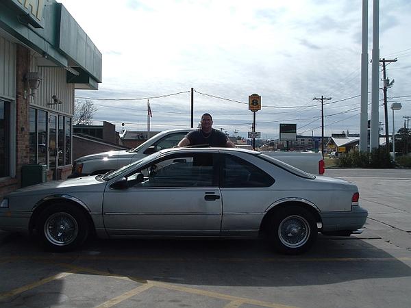 Silver Ford Thunderbird coupe parked in front of storefront, man standing behind it