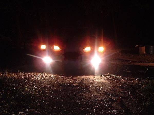 Front of a dark car at night with headlights and fog lights glowing over a dirt yard