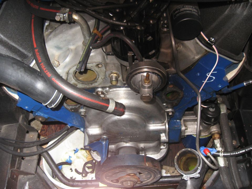Close-up of a V8 engine, showing hoses, distributor, oil filter, and pulleys. Blue painted brackets are visible