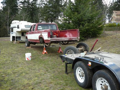 1991 Ford Bronco - A red and white Ford pickup truck is raised on jack stands in a grassy yard, with its rear axle removed and resting on a trailer. A camper…