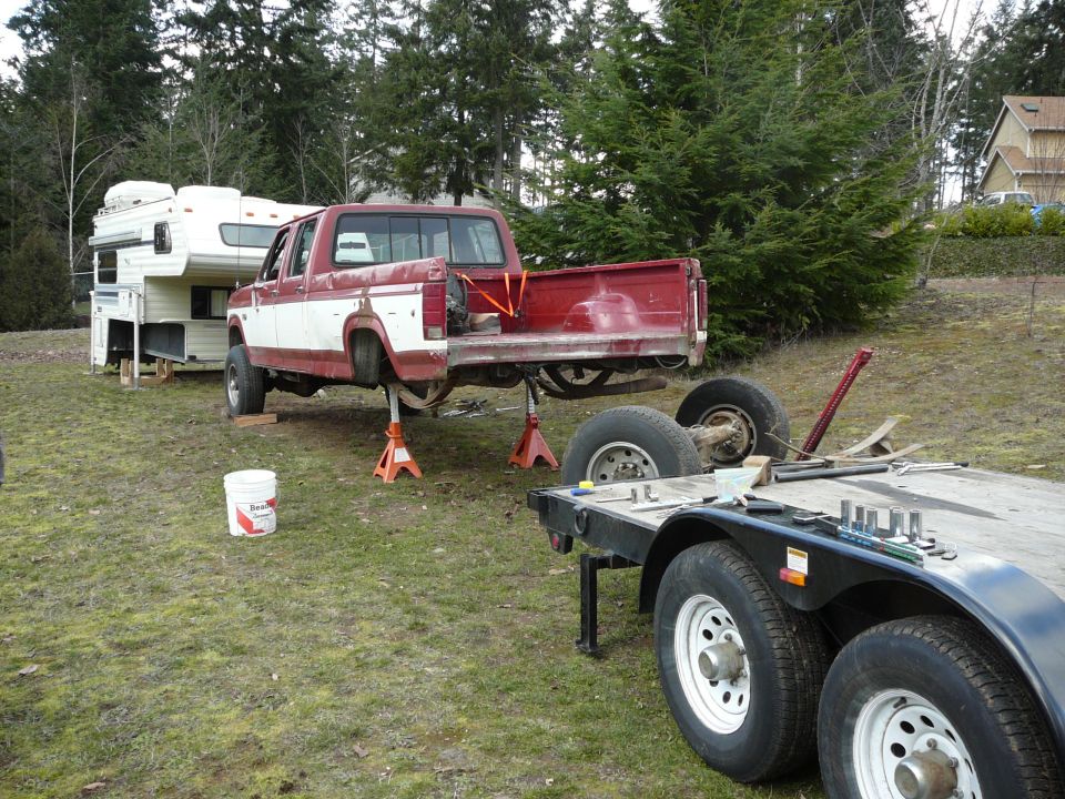 Red and white Ford pickup truck on jack stands with rear axle removed, camper shell in background.