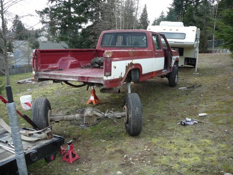 1991 Ford Bronco - A red and white Ford pickup truck is raised on jack stands with its rear axle removed and resting on a trailer. A white camper is parked be…