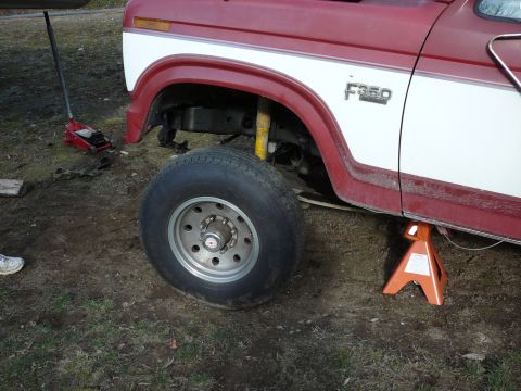 1991 Ford Bronco - A red and white Ford F350 truck is jacked up with a jack stand under the frame and the front wheel removed. A red hydraulic floor jack is v…