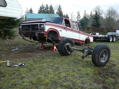1991 Ford Bronco - A red and white pickup truck is on jack stands with its front end elevated and rear axle removed. Tools and a trailer are visible in the ba…