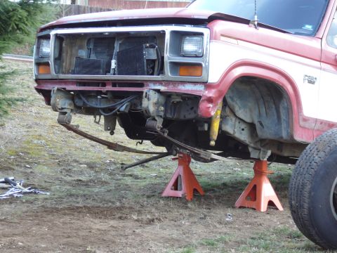 1991 Ford Bronco - A red and white Ford truck is elevated on jack stands, with its front end disassembled. The grille is missing, and the front bumper and lea…