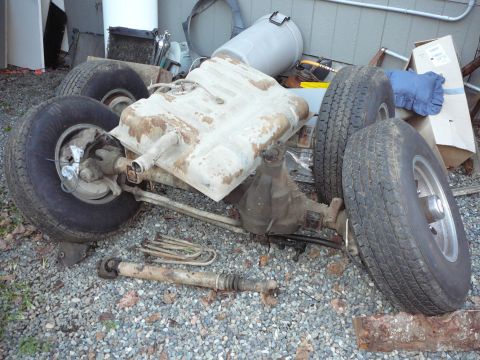 1991 Ford Bronco - A disassembled rear axle assembly with two tires, a fuel tank, and a driveshaft is laid out on a gravel surface.