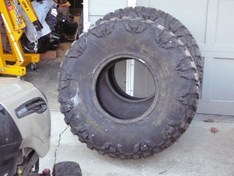 1991 Ford Bronco - Two large, aggressive off-road tires are leaning against a garage door. A portion of a tan riding mower is visible in the foreground to the…