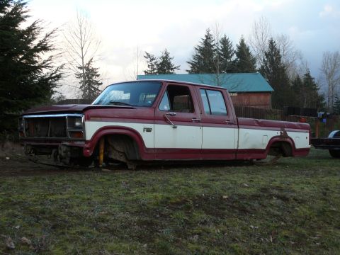 1991 Ford Bronco - A maroon and white pickup truck, missing its wheels and bed, sits on a grassy field. The front of the truck is visible, showing a damaged g…