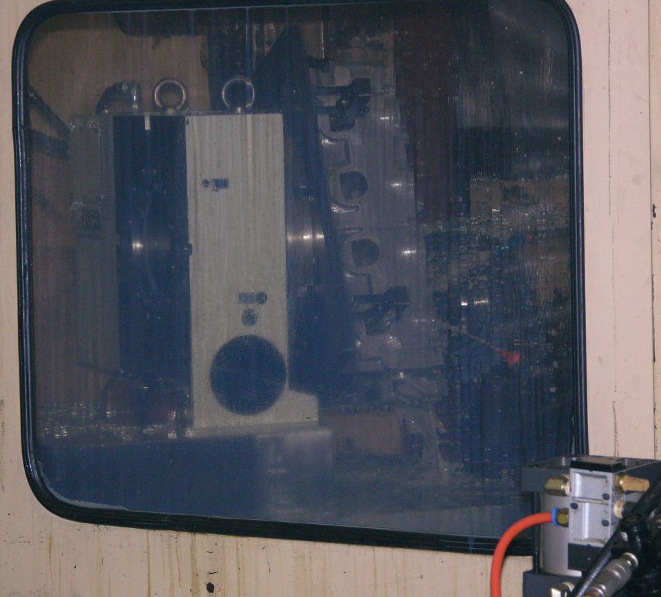 Cylinder head fixtured inside a CNC machine, seen through a coolant-spattered window