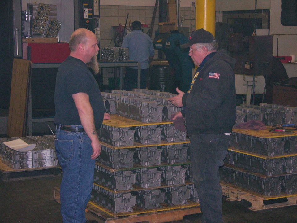 Two men talking beside pallets of aluminum cylinder head castings in a machine shop