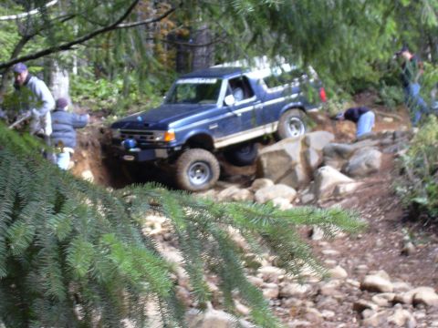1991 Ford Bronco - A blue 1991 Ford Bronco is navigating a rocky, wooded trail. Several people are visible around the vehicle, assisting or observing.
