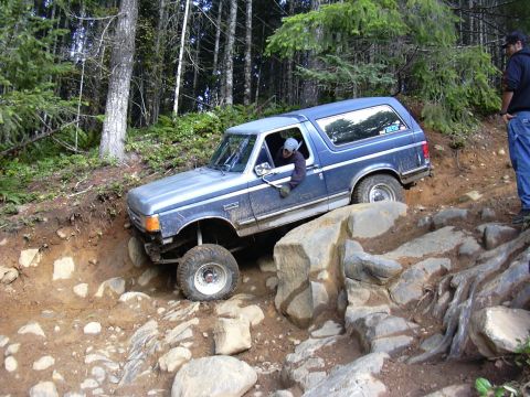 1991 Ford Bronco - A blue 1991 Ford Bronco is navigating a steep, rocky, and muddy off-road trail in a forest. The driver is visible in the driver's seat, and…