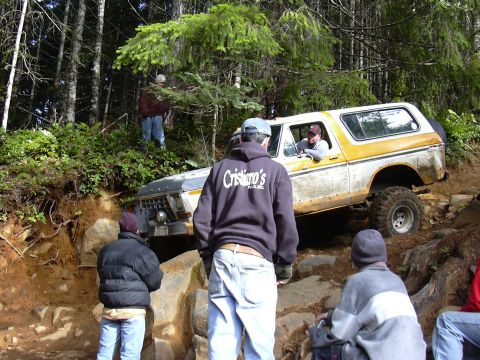 1991 Ford Bronco - A yellow and white Ford Bronco is navigating a steep, rocky incline in a wooded area. Several people are standing around the vehicle, some…