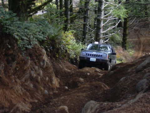 1991 Ford Bronco - A dark green Jeep Grand Cherokee is driving off-road on a dirt trail through a forest. The vehicle is navigating a steep, rutted path with…