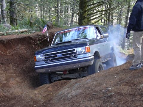 1991 Ford Bronco - A dark blue and silver Ford Bronco is navigating a steep dirt ditch with smoke coming from the tires. An American flag is mounted on a pole…