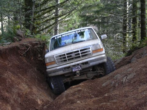 1991 Ford Bronco - A white 1990s Ford Bronco is navigating a steep, rutted dirt trail in a forest. The vehicle is tilted at a sharp angle as it climbs out of…