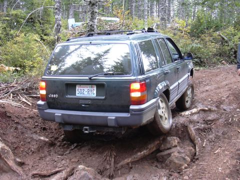 1991 Ford Bronco - A dark green Jeep Grand Cherokee is driving off-road through mud and rocks on a wooded trail. The vehicle's rear is angled towards the came…