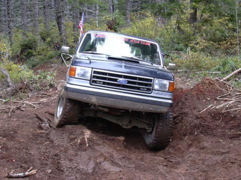 1991 Ford Bronco - A dark blue Ford Bronco is navigating a steep, muddy, off-road trail with large tires and an American flag mounted on the windshield frame.…