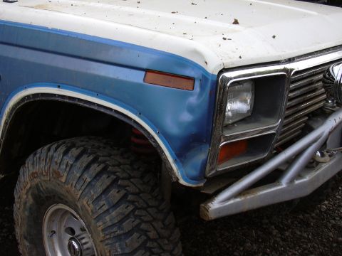 1991 Ford Bronco - Close-up of the front driver's side fender, headlight, and tire of a blue and white off-road vehicle. A custom metal bumper is visible belo…