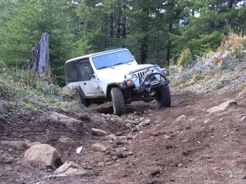 1991 Ford Bronco - A white Jeep Wrangler is navigating a rocky, uphill trail in a wooded area. The off-road vehicle is tilted to the side as it climbs over ob…