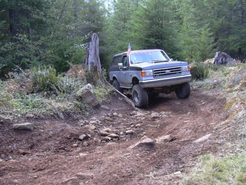 1991 Ford Bronco - A two-tone grey and blue Ford Bronco is driving off-road up a dirt trail in a wooded area. The vehicle has large tires and an American flag…
