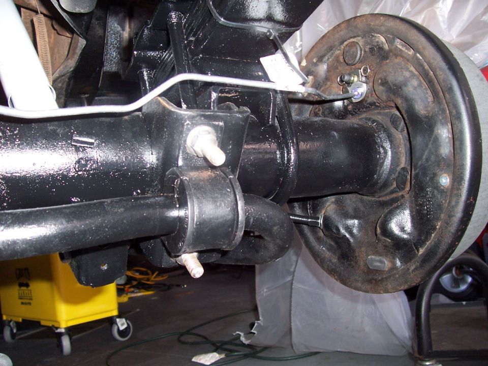 Rear axle assembly with brake drum and suspension components. A yellow bucket is visible in the background